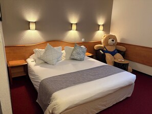 Una cama bien hecha con una manta gris, dos almohadas estampadas y un osito de peluche sobre una mesita redonda.