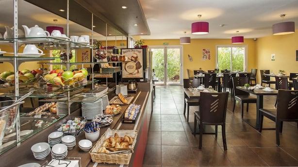 Uma área de buffet com diversos alimentos, incluindo frutas, doces e bebidas. Há várias mesas e cadeiras dispostas para os hóspedes.