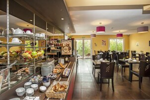 Uma área de buffet com diversos alimentos, incluindo frutas, doces e bebidas. Há várias mesas e cadeiras dispostas para os hóspedes.