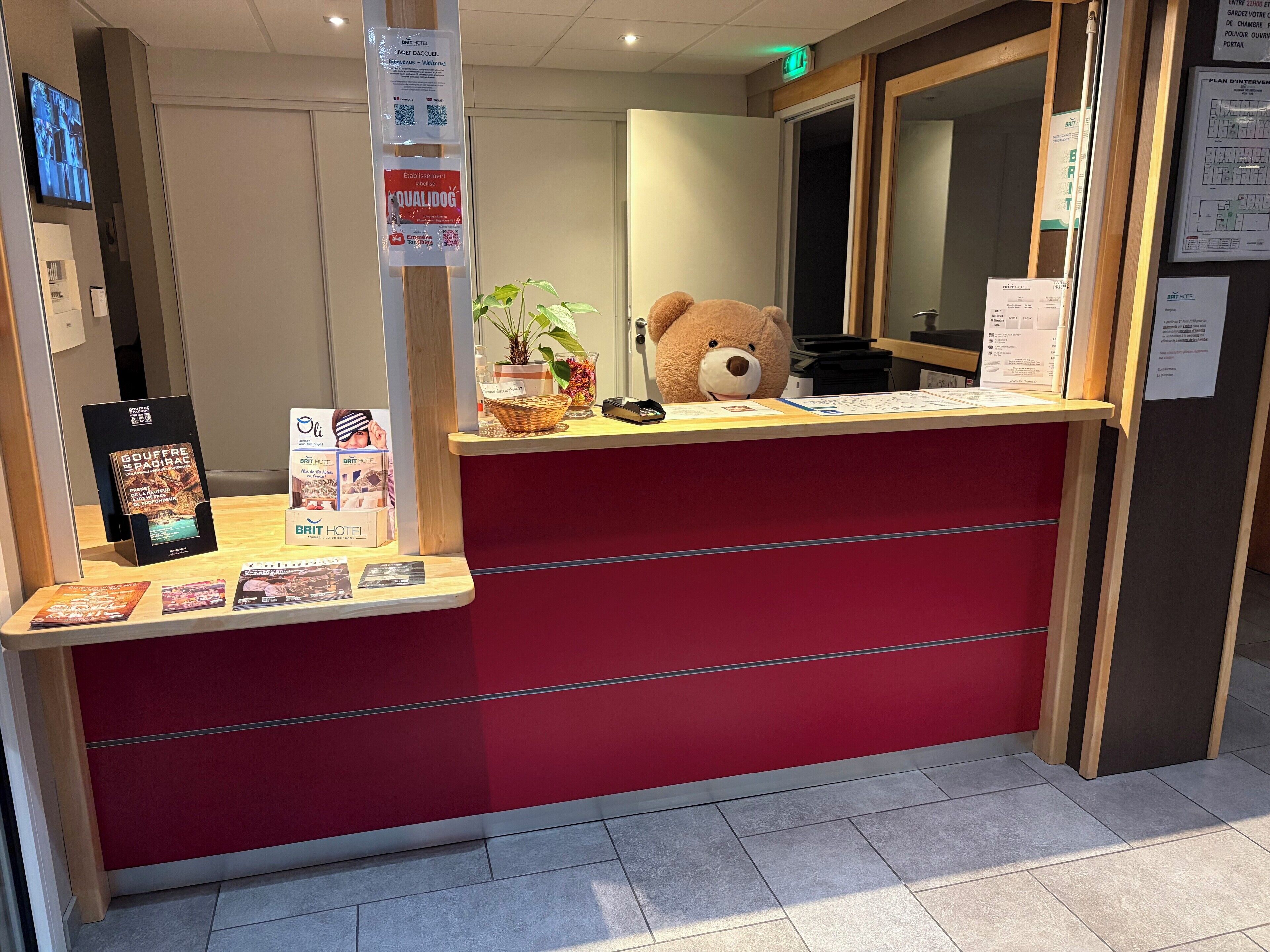 Bancada de recepção com superfície vermelha, um urso de pelúcia grande e cartazes informativos.