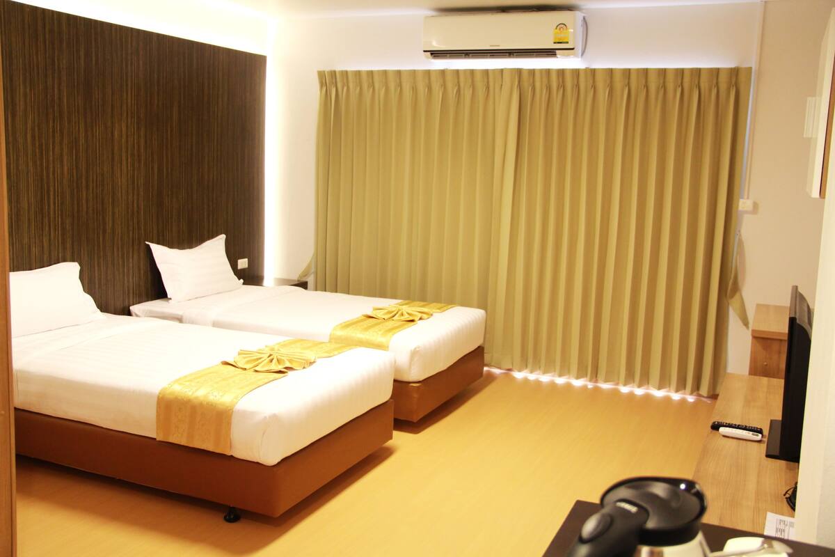 A hotel room with two beds, a wooden headboard, a large window with curtains, an air conditioning unit, and a television.