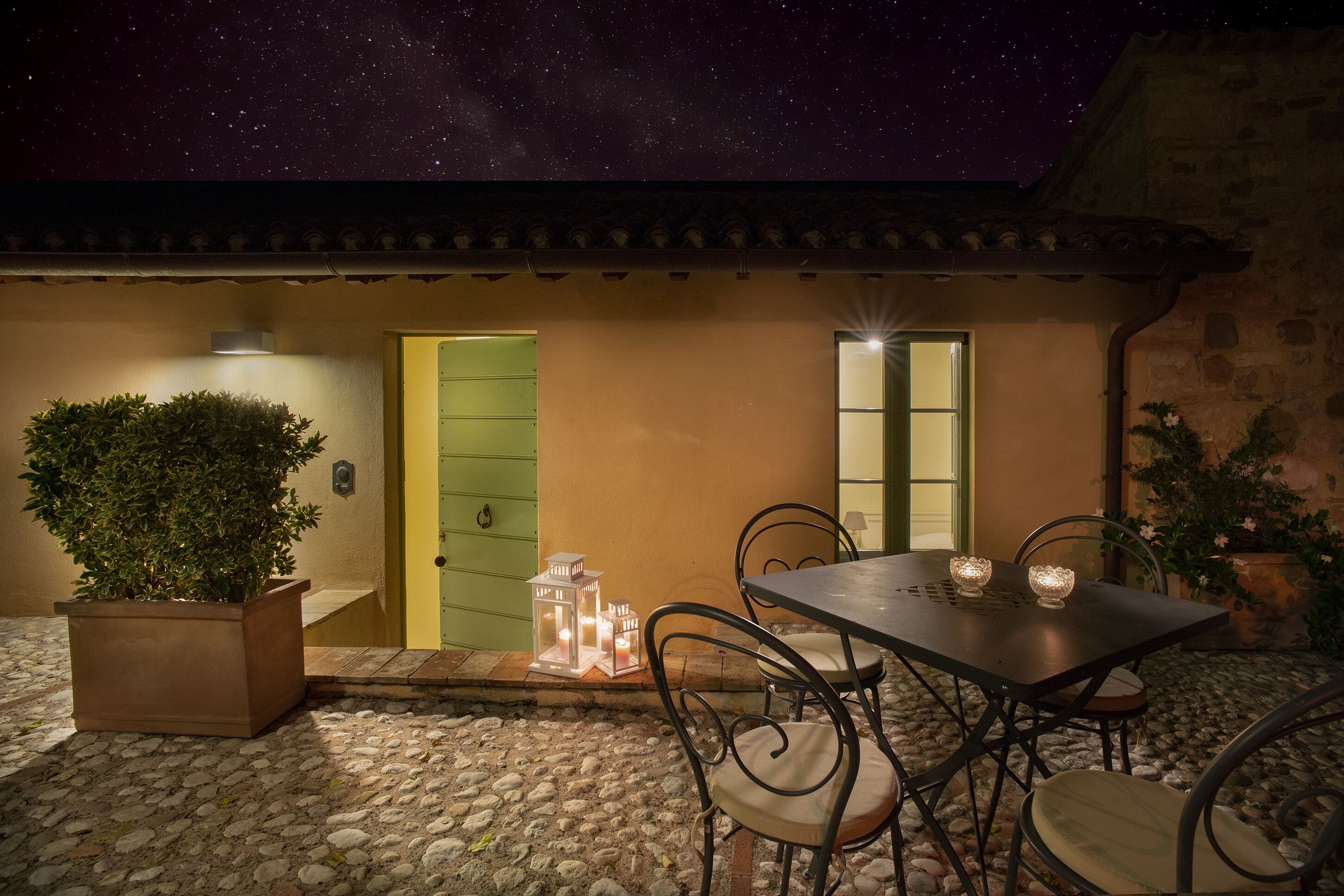 An outdoor patio with a table and chairs, a potted plant, and a door with a light.