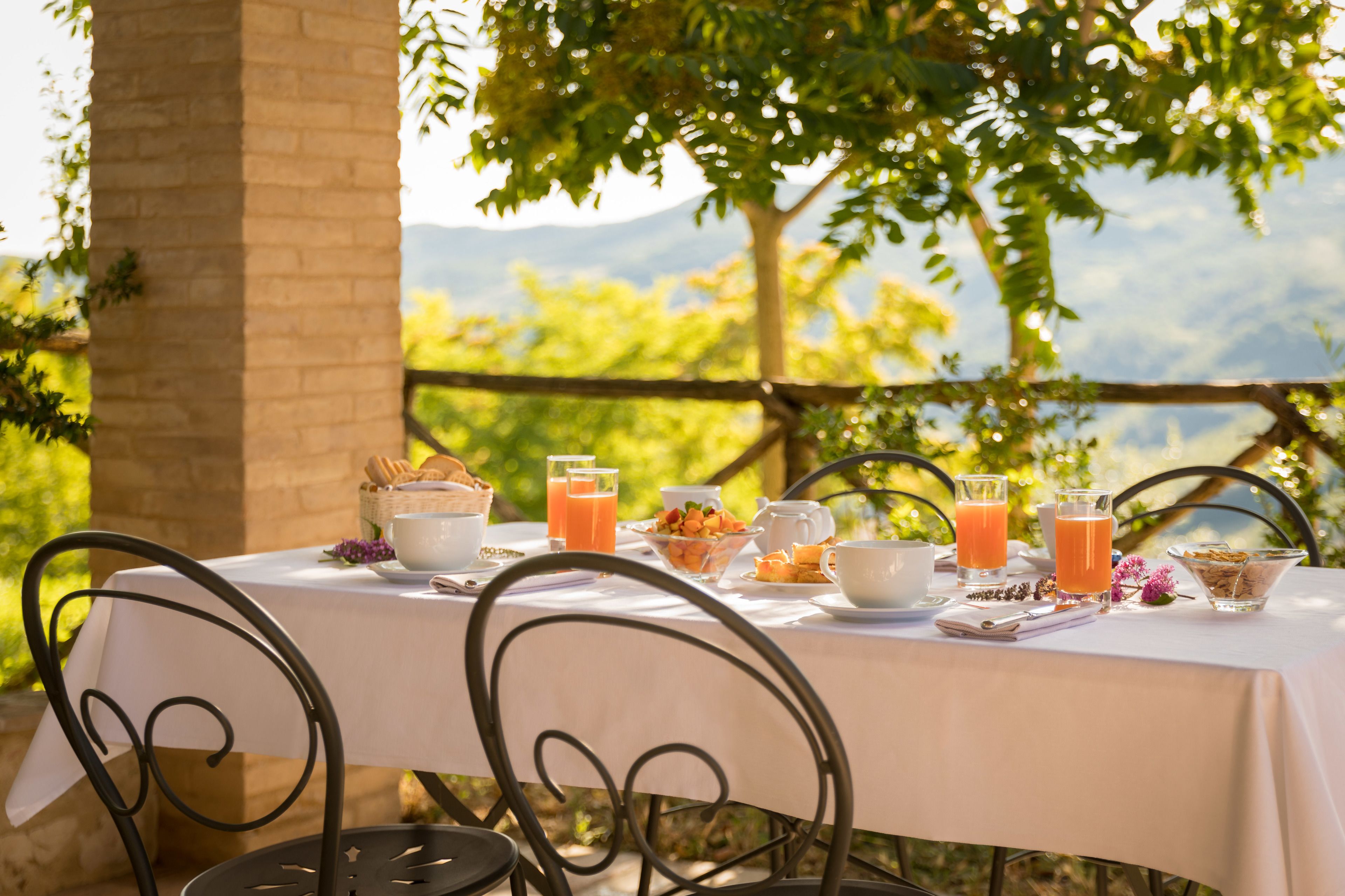 A table set outdoors with breakfast items, including bread, fruit, and beverages, surrounded by chairs.