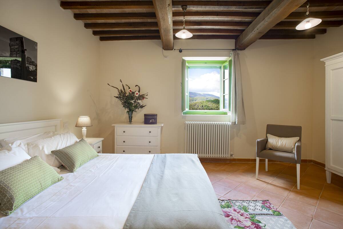 A bedroom with a bed, a chair, a dresser, a window with a view, and a radiator.