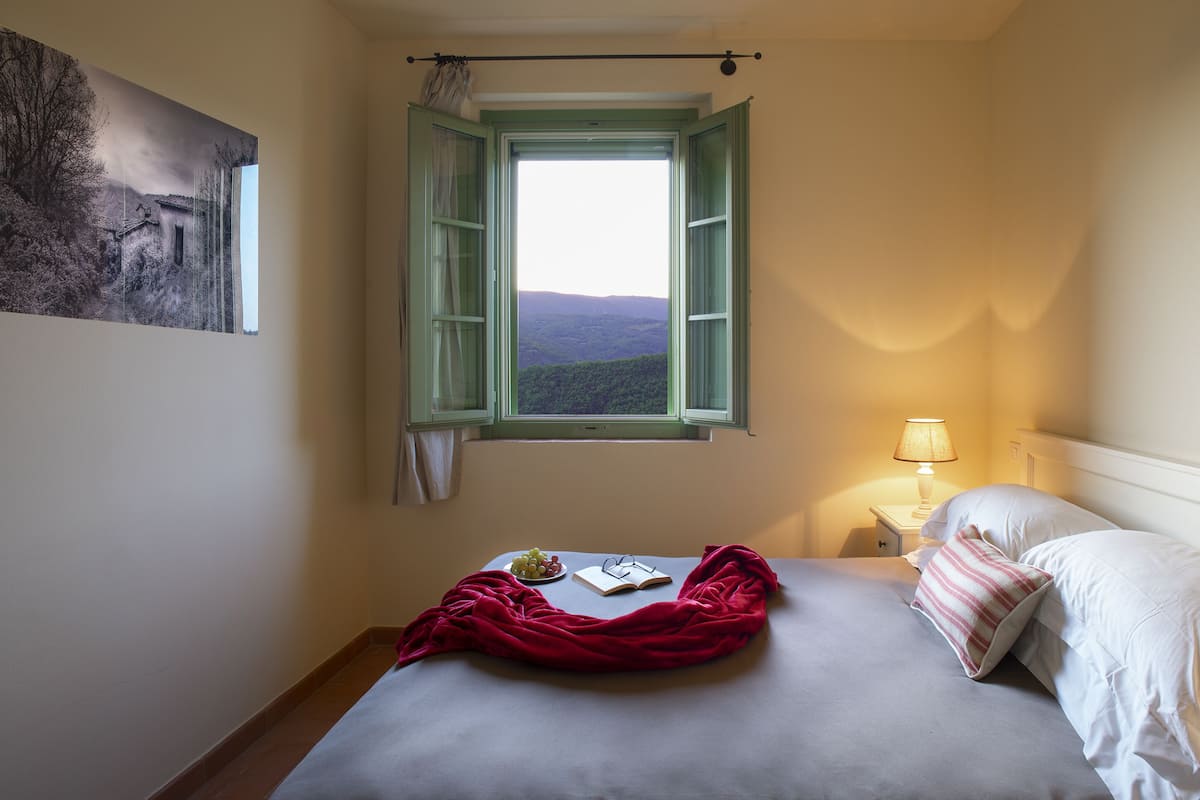 A bedroom with a bed, a window with open shutters, a bedside table with a lamp, and a painting on the wall.