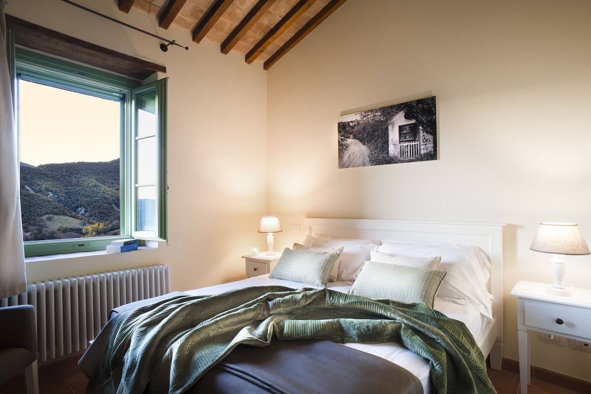 A bedroom with a bed, a window with a view of hills, a bedside table with a lamp, and a wall-mounted picture.