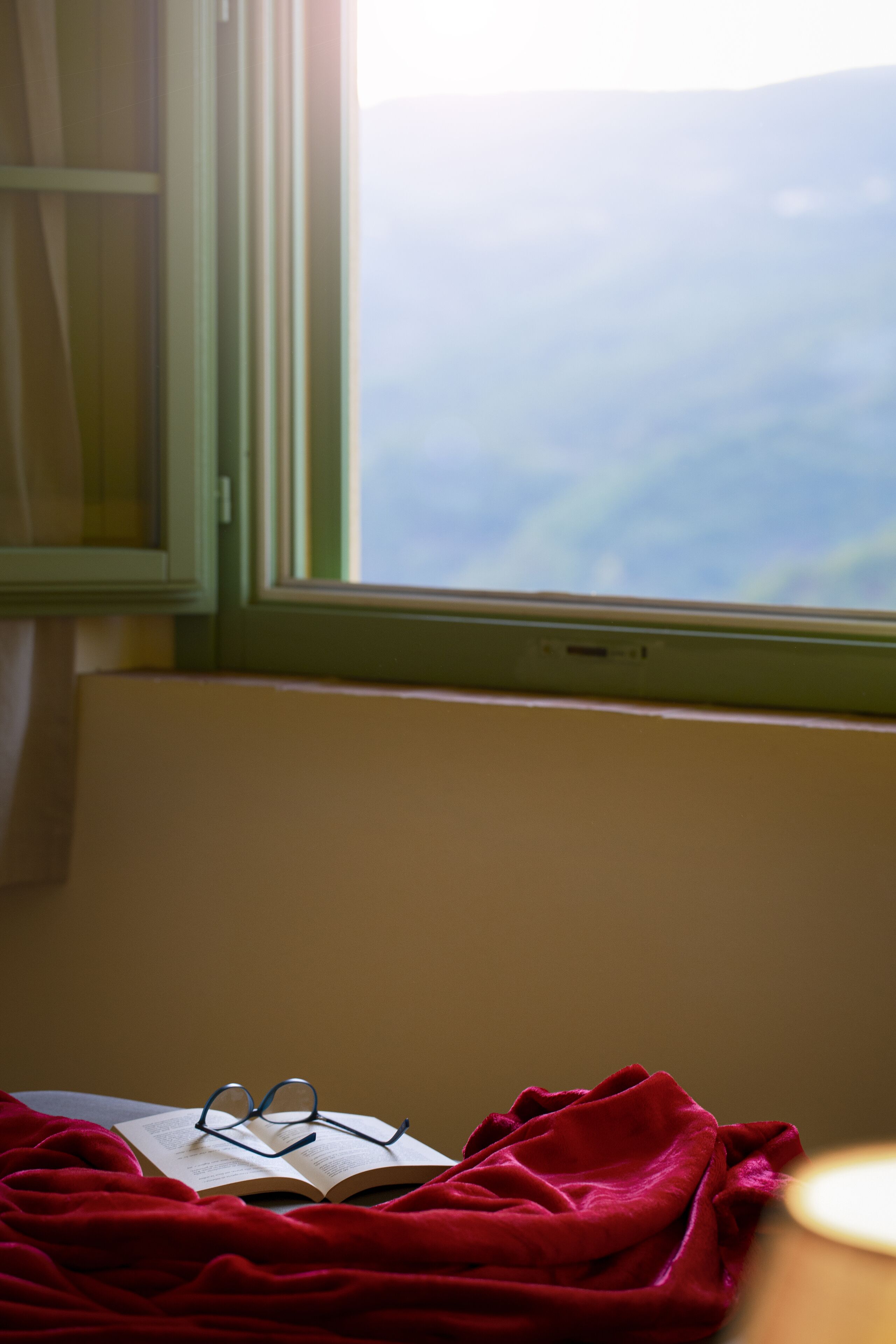 Una stanza con una finestra, un letto con una coperta rossa e un paio di occhiali su un libro aperto.
