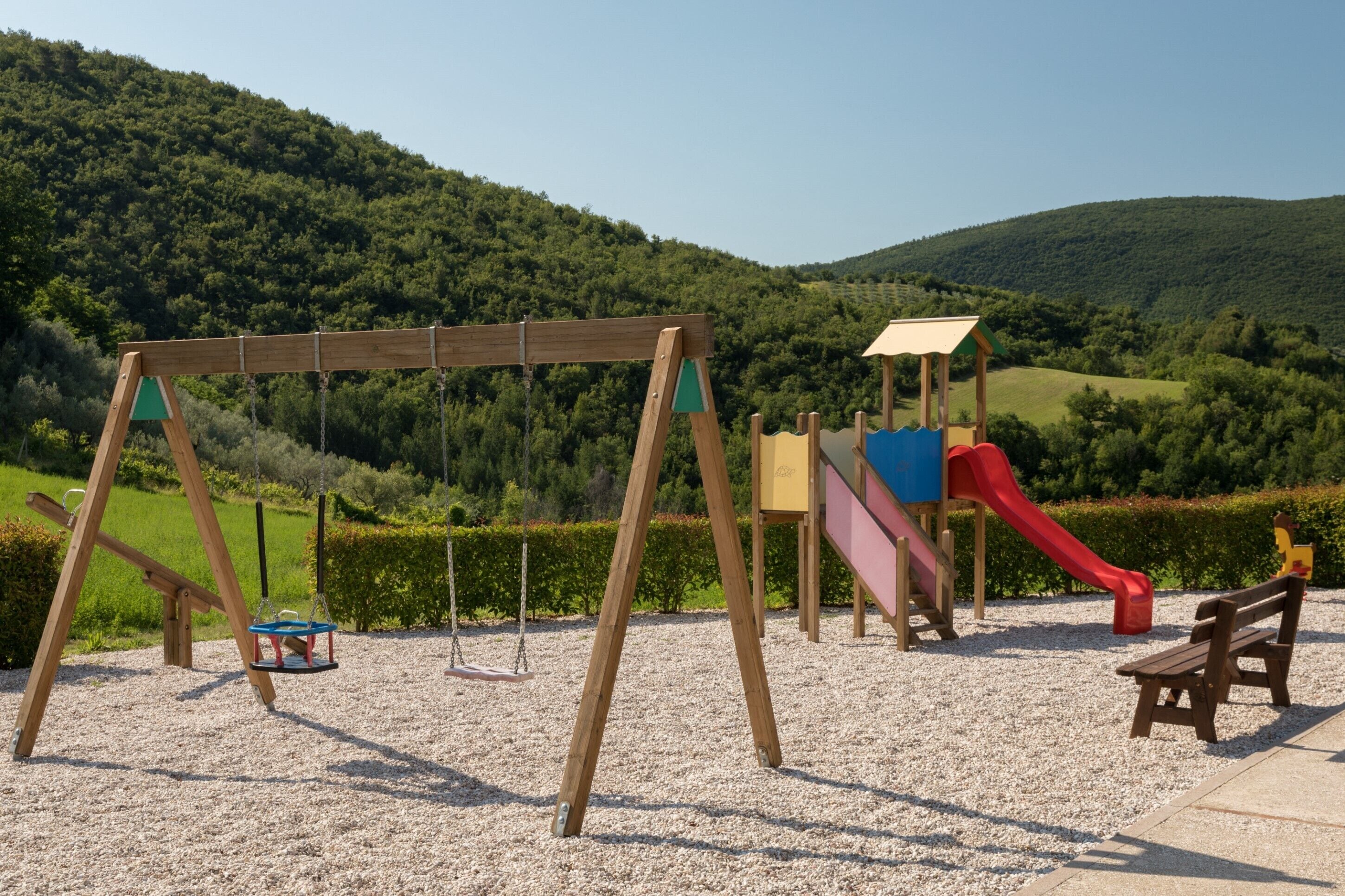 A playground with swings, a slide, and a bench in a park with a hilly backdrop.