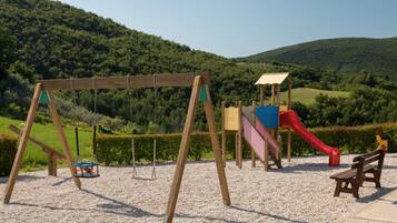 A playground with swings, a slide, and a bench in a park with a hilly backdrop.