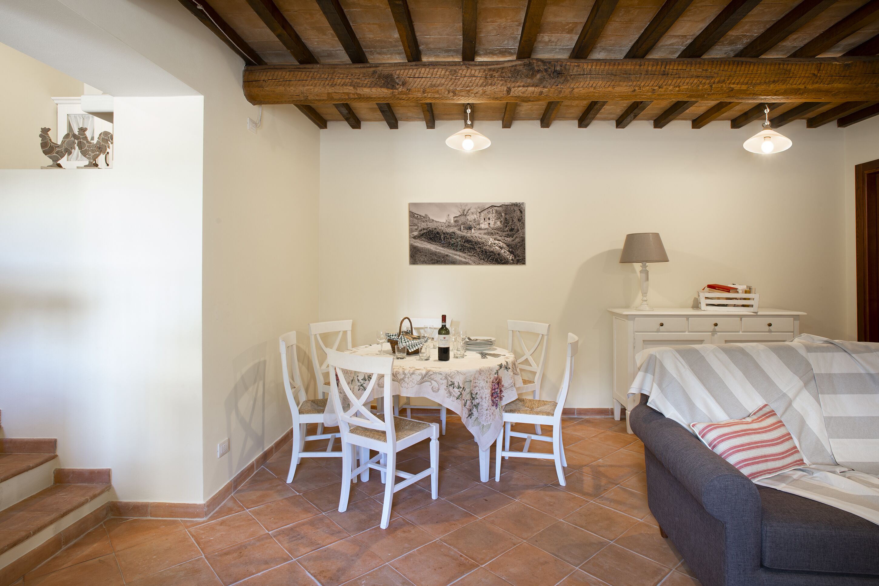 A dining area with a table set for four, white chairs, a painting on the wall, and a sofa with a striped blanket.