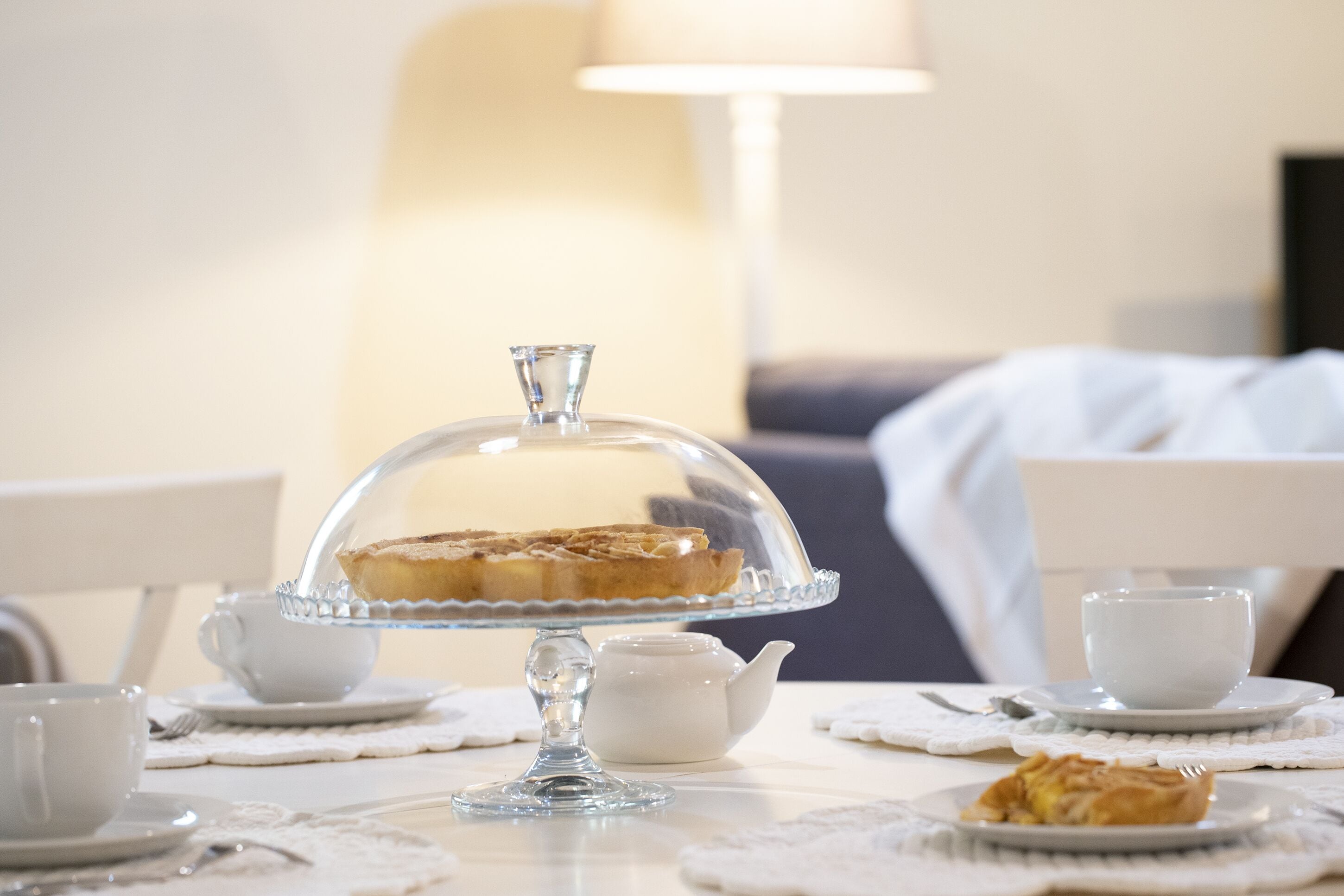 Una mesa de comedor con un pastel cubierto, una tetera y tazas sobre un mantel blanco.