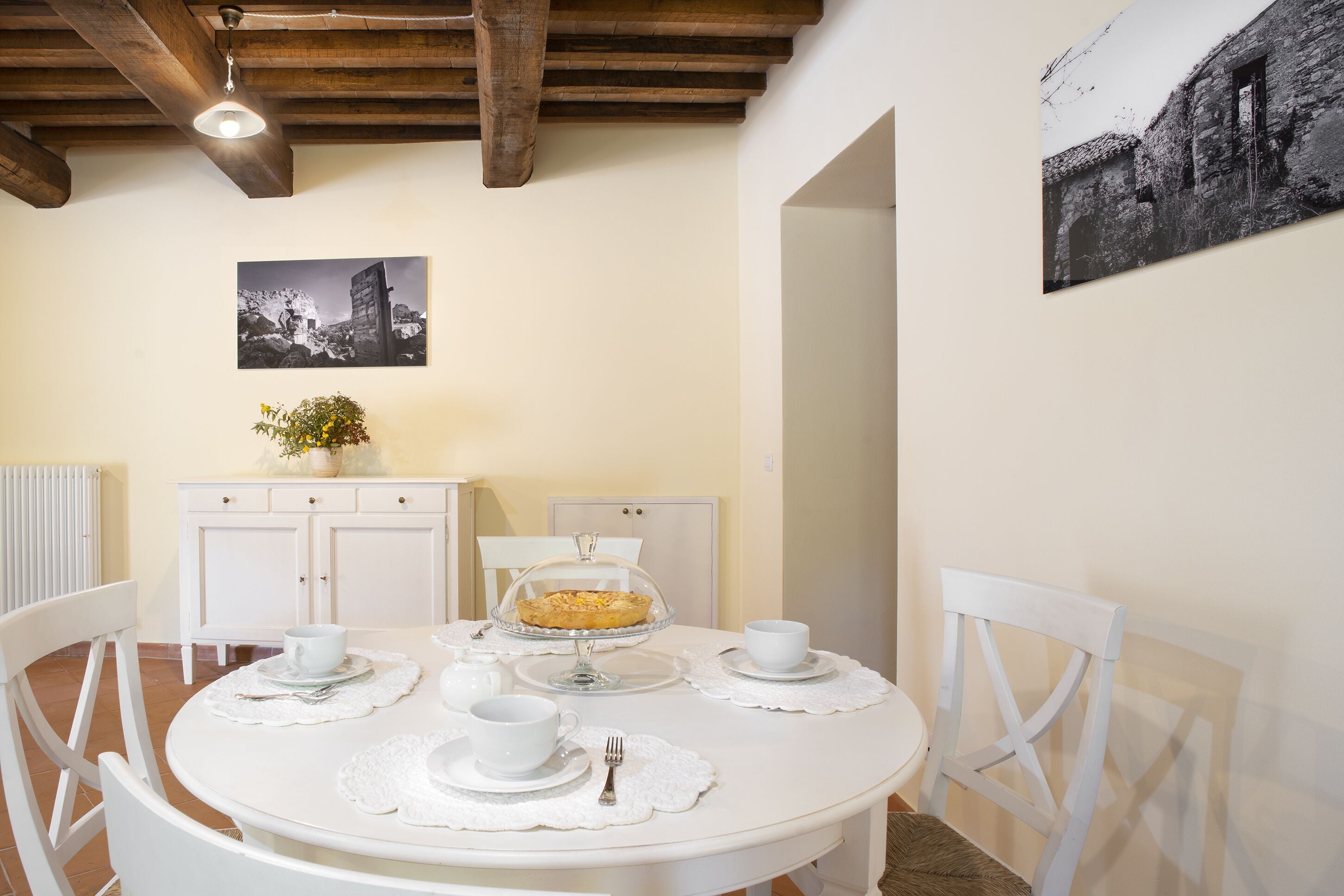 A dining area with a round table set for four, white chairs, a white cabinet with a plant, and a black and white photograph on the wall.