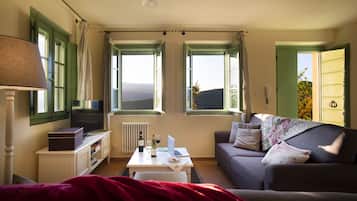 A cozy living room with a TV, laptop, and wine glasses on a table, a sofa with cushions, and large windows with curtains.