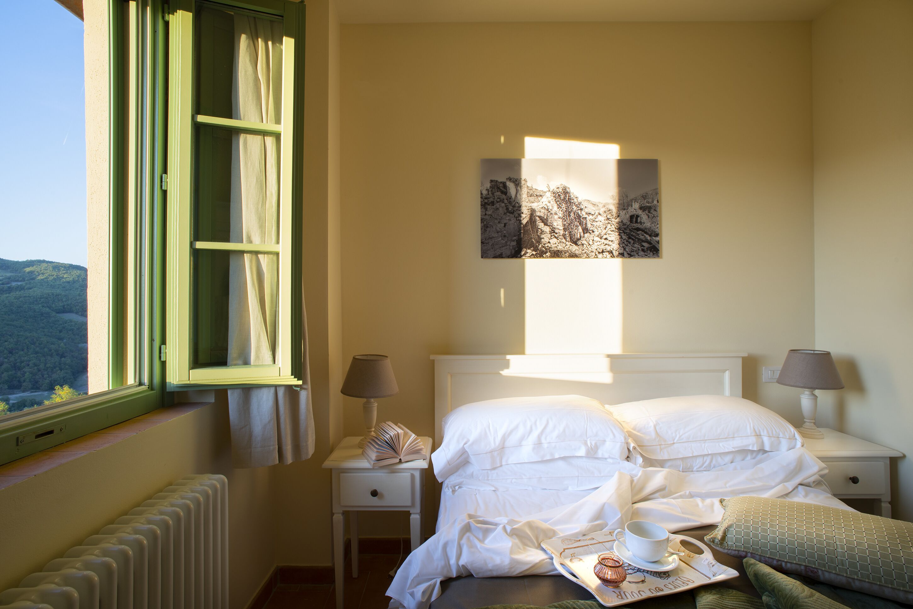 Habitación de hotel con cama, dos mesitas de noche, ventana con cortinas, radiador y un cuadro en la pared.