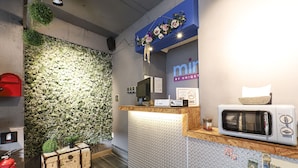 Ein Empfangsbereich mit blauer Wand, einer Wandpflanze, einem Empfschalter, einem Computer, einer Mikrowelle und einem roten Briefkasten.