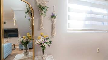 Una habitación con un espejo grande, dos adornos de macramé colgados en la pared con plantas, y una mesa con un jarrón de flores.