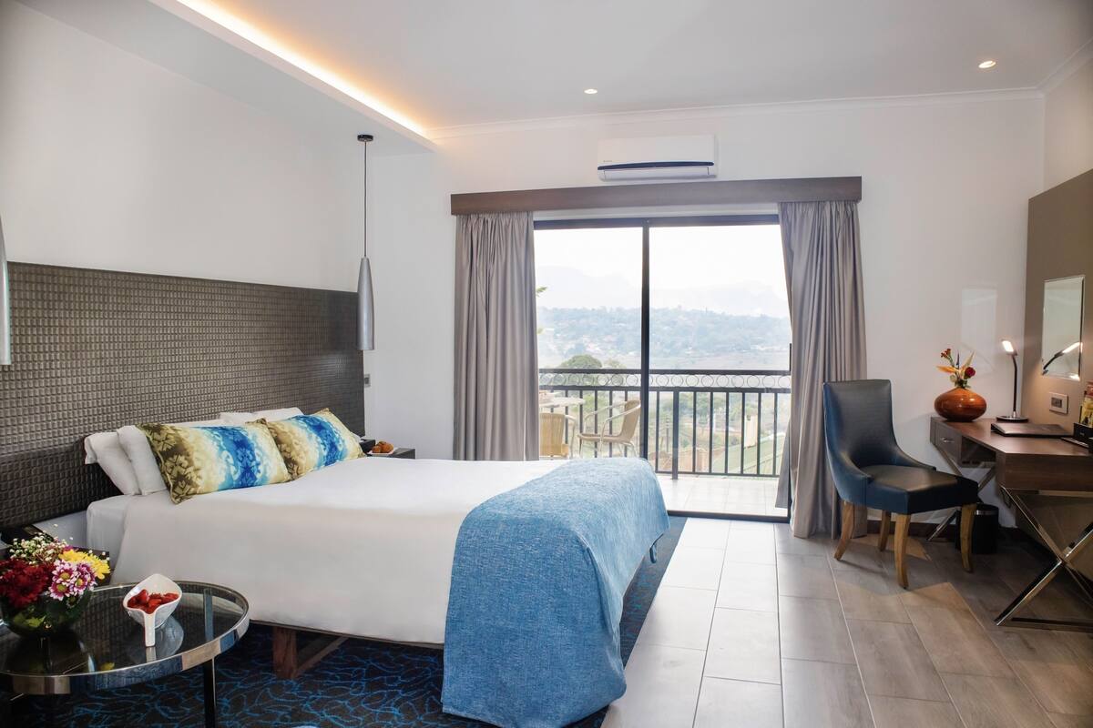 A hotel room with a bed, a desk, a chair, a balcony with a view, and a dining area.