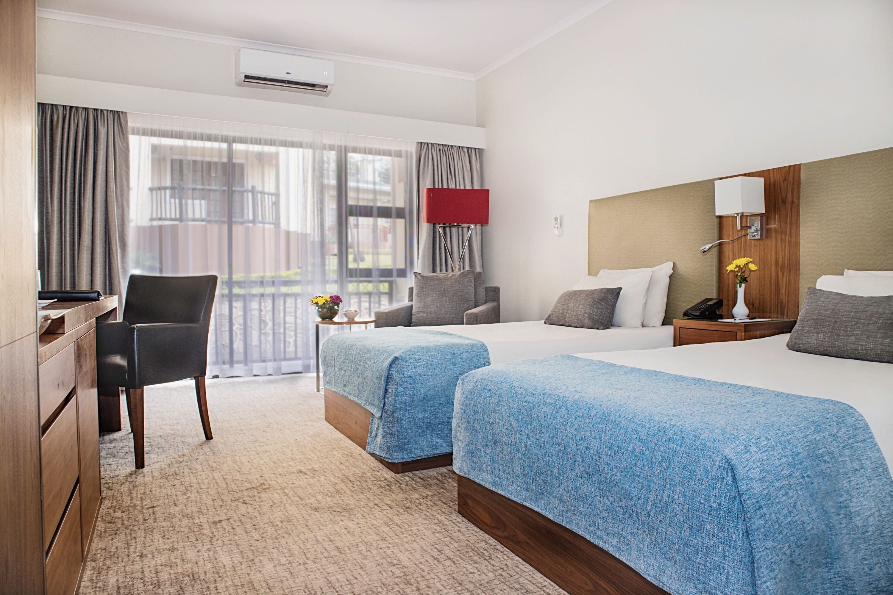 A hotel room with two beds, a desk, a chair, a lamp, and a view of the outdoors.