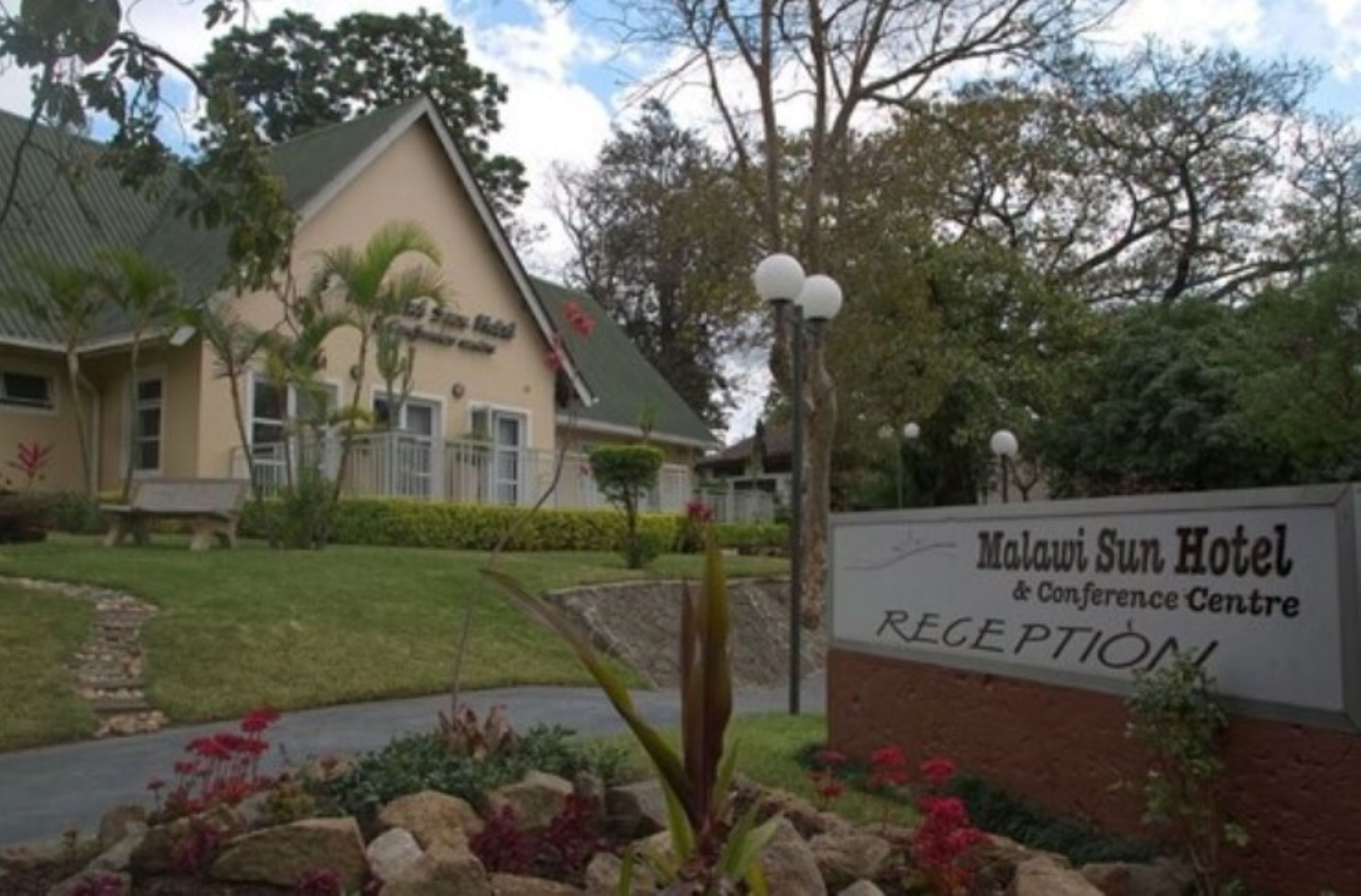 The Malawi Sun Hotel & Conference Centre reception area with a sign, a building, and surrounding greenery.