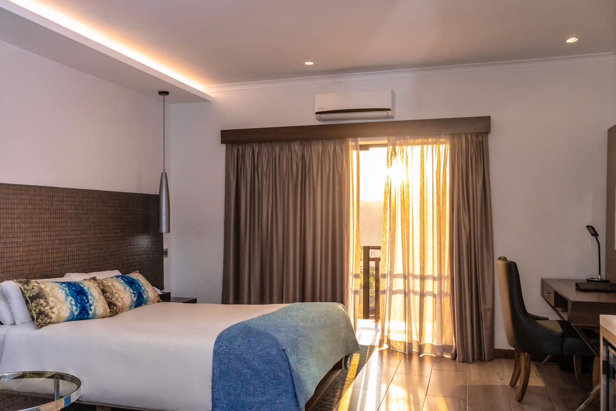 A hotel room with a bed, a desk, a chair, an air conditioning unit, and a view of the outdoors through a window with curtains.
