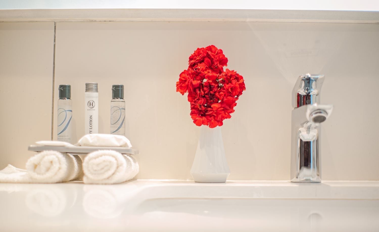 A bathroom with a sink, a vase with red flowers, and toiletries.