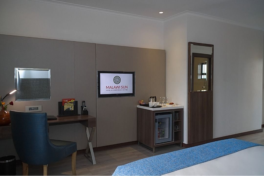 A hotel room with a desk, a television displaying 'MALAWISUN HOTEL & CONFERENCE CENTRE', a wardrobe, and a bed with a blue bedspread.