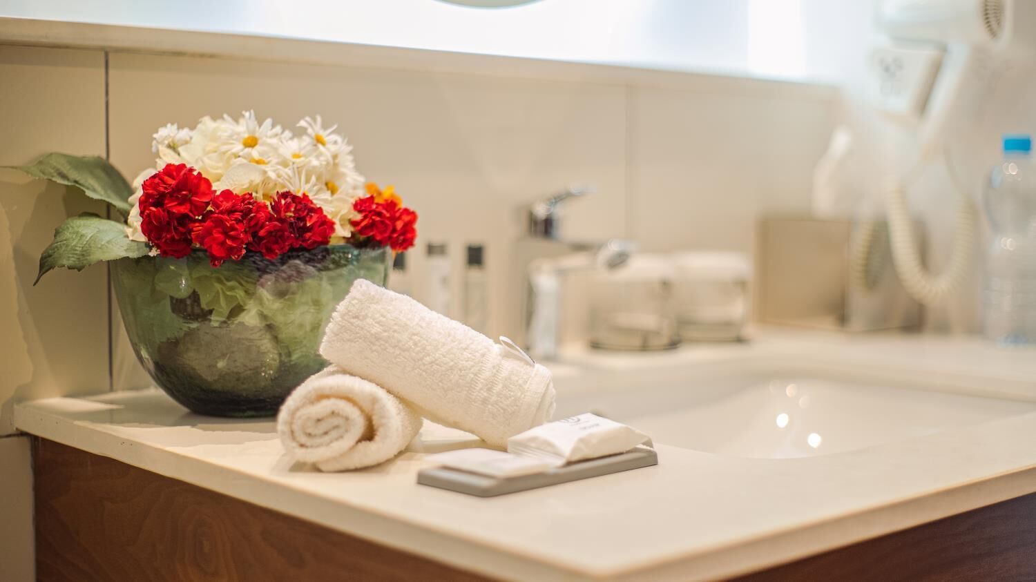A bathroom with a glass vase containing red and white flowers, a rolled white towel, and toiletries on the counter.