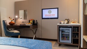 Quarto de hotel com cama, escrivaninha com computador, televisão exibindo 'Malawi Sun Hotel & Conference Centre', frigobar e uma cadeira azul.