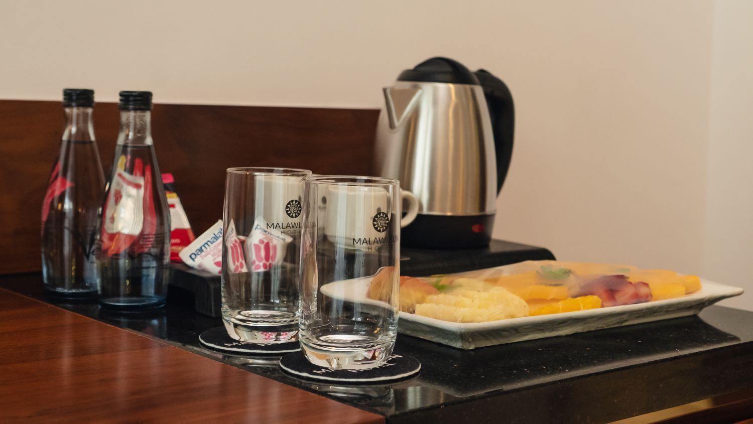 Une table dans une chambre d’hôtel avec deux verres, une bouteille, une bouilloire et une assiette de nourriture.