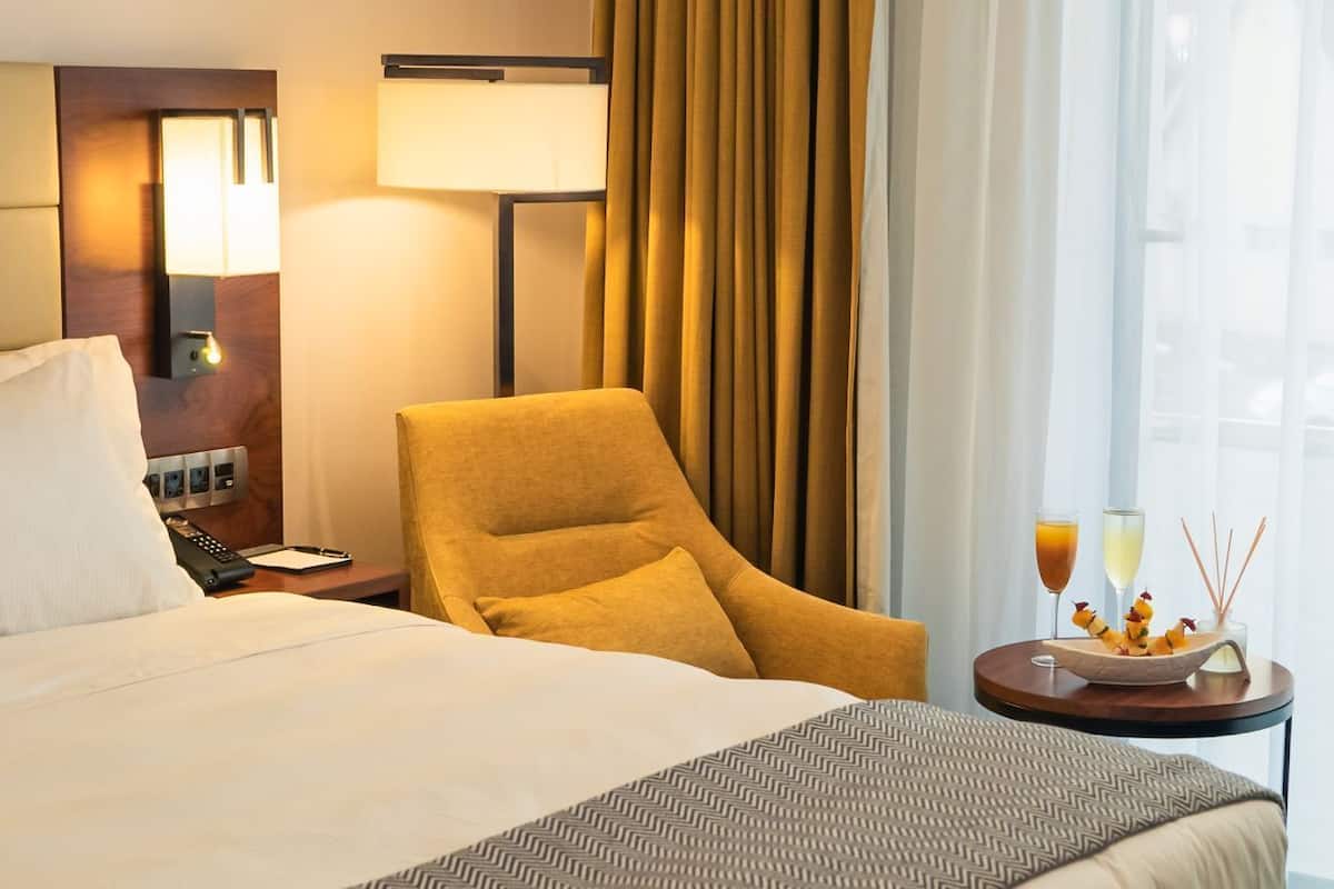 A hotel room with a bed, a chair, a nightstand with a lamp, a table with a drink and snacks, and a window with curtains.