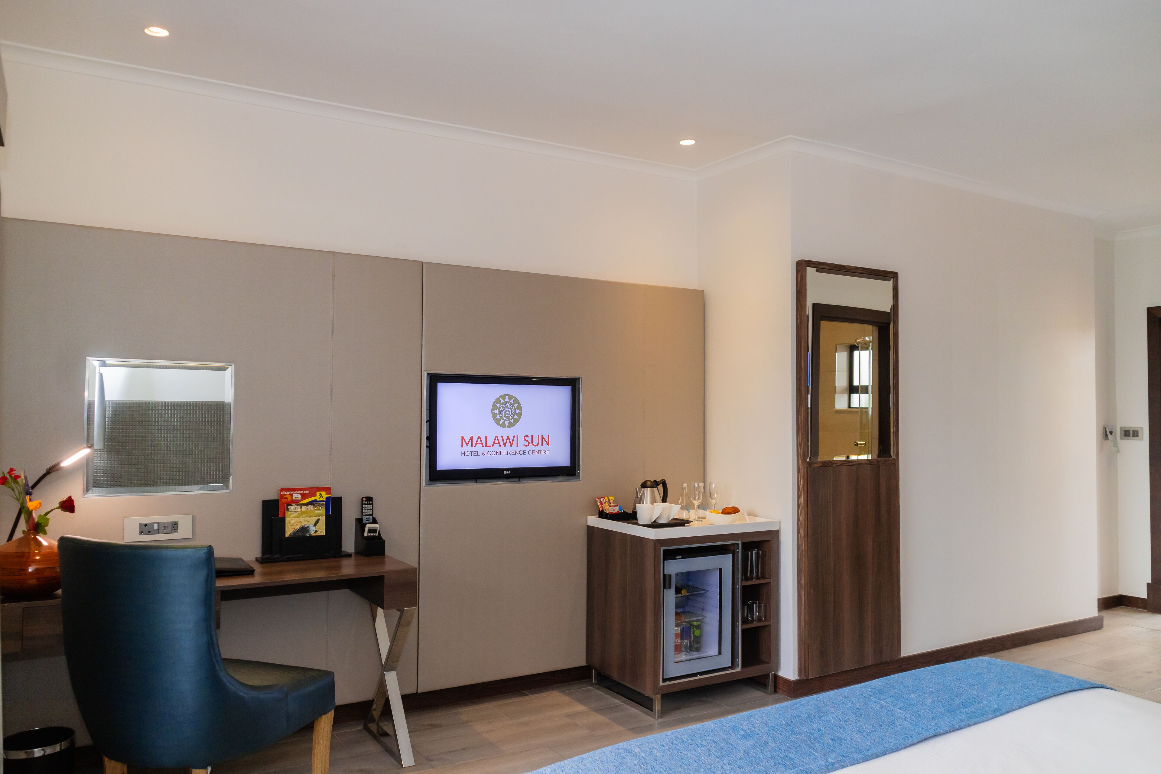 Una habitació d'hotel amb un llit, una taula de nit amb un ordinador, un televisor que mostra 'Malawi Sun Hotel & Conference Centre', un armari i una cuineta petita.