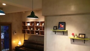 A living room with a brown leather sofa, a green coffee table, and a hanging green pendant light. There are shelves with framed pictures and a small plant on a yellow shelf. - Popcorn Hostel Nampodong (Busan)