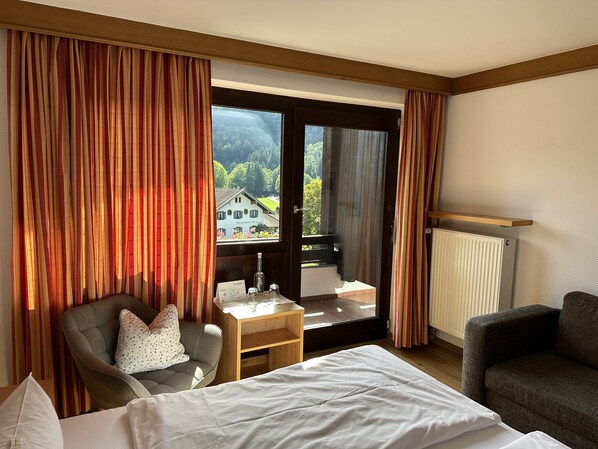 A hotel room with a bed, a chair, a small table with a bottle and glasses, and a view of a building through a large window with curtains. - Landhotel Maiergschwendt by DEVA (Ruhpolding)