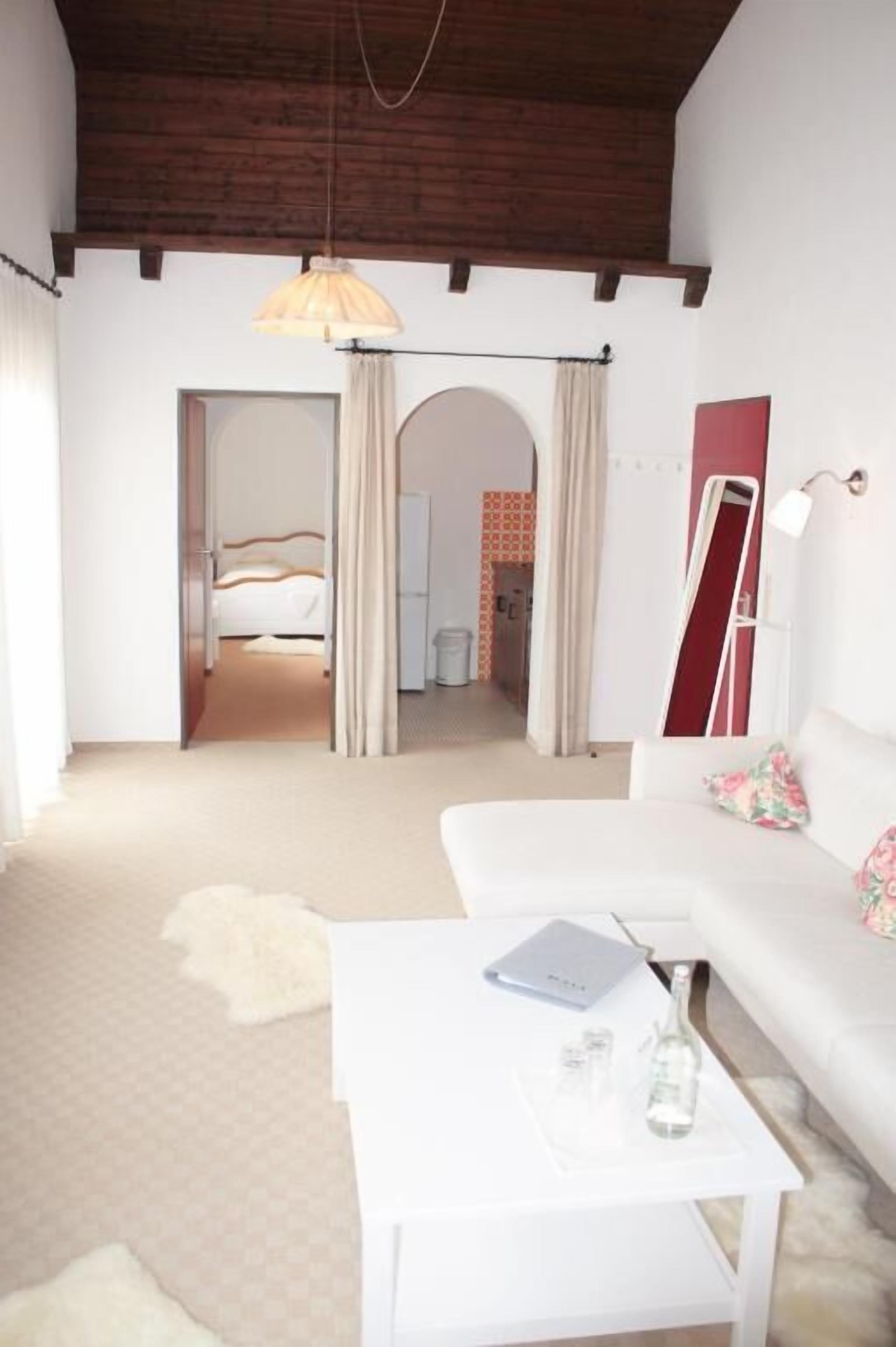 Ein Zimmer mit einem weißen Sofa, einem kleinen weißen Tisch mit einem Laptop und einer Flasche, einer Hängelampe und einem Bett im Hintergrund.