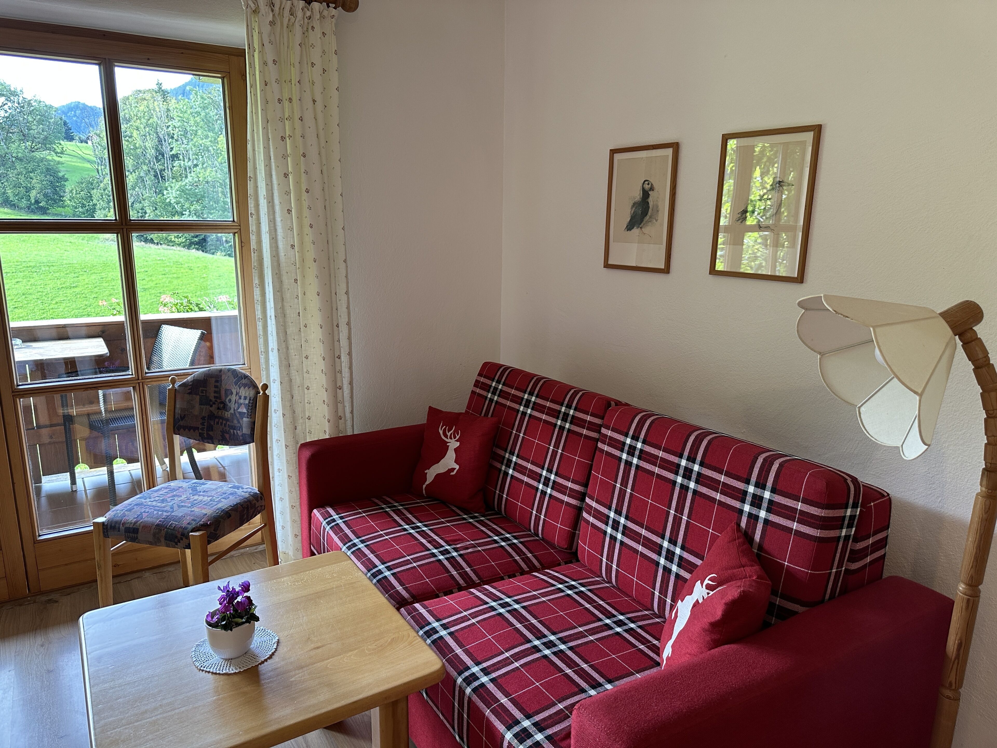 A cozy living room with a red plaid sofa, a wooden table with a potted plant, and a view of a green landscape through a large window.