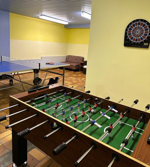 Una mesa de fútbol de mesa con un dardo y una mesa de ping pong en una habitación con paredes amarillas y piso de baldosas.