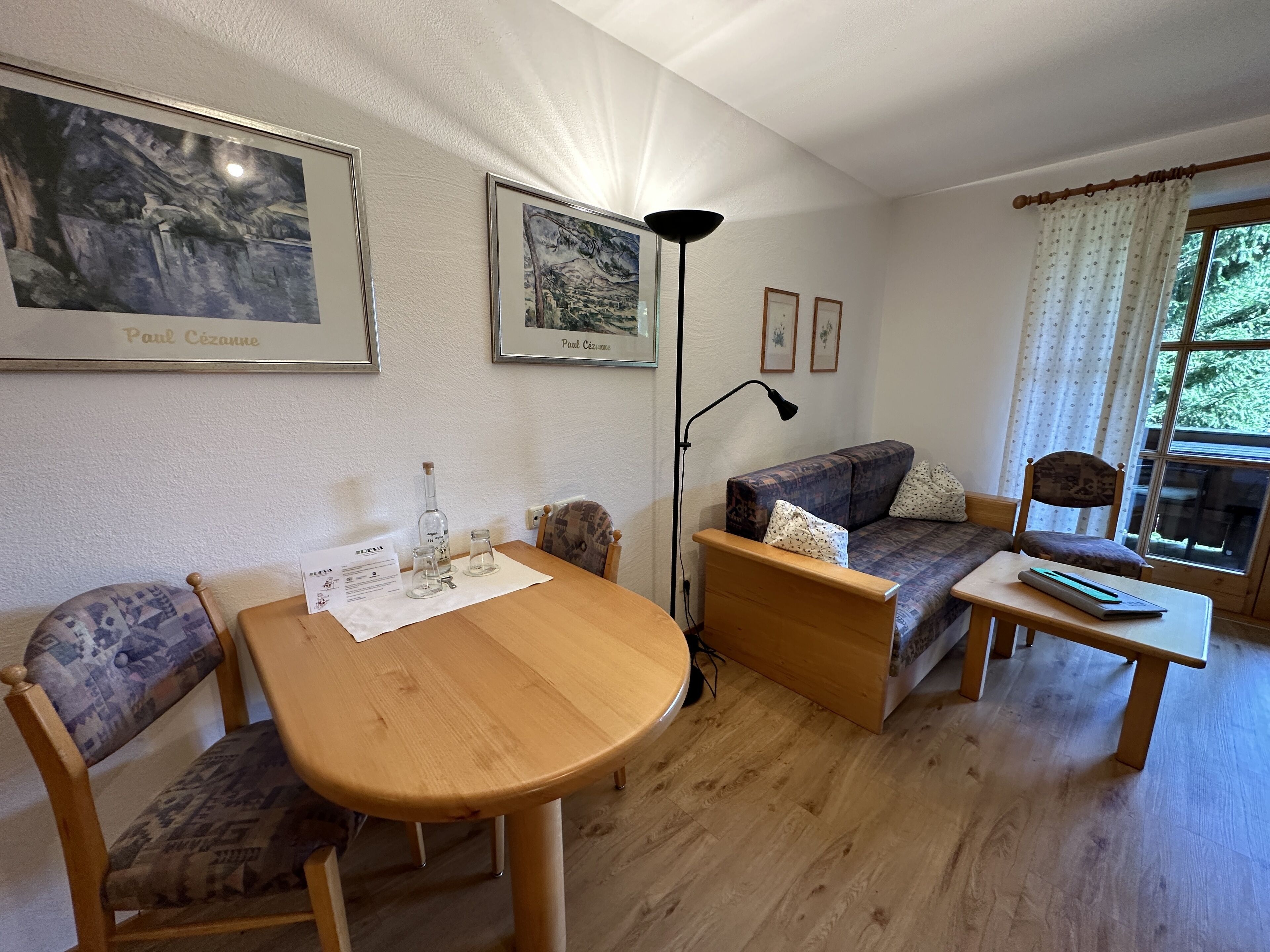 A cozy living room with a wooden table, two patterned chairs, a sofa, and a small side table. There are framed artworks on the wall, a floor lamp, and a window with curtains.