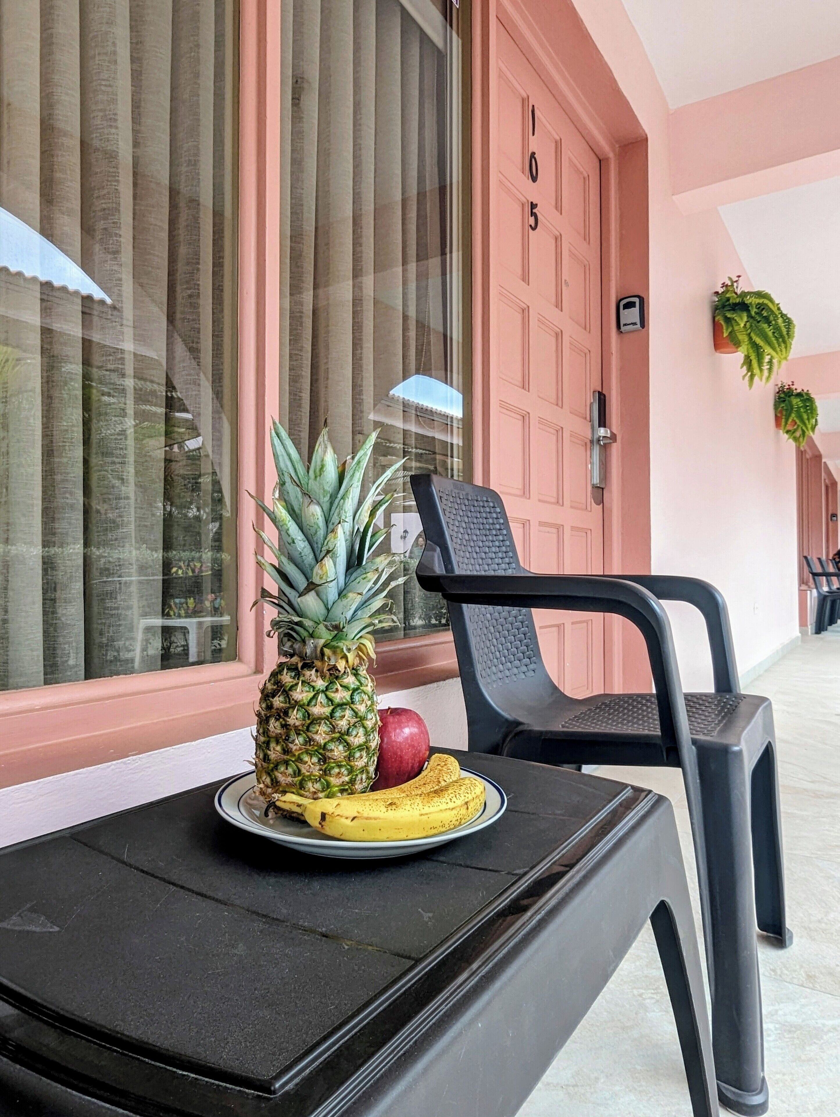Una mesa con una fuente de piña, plátano y manzana, una silla negra y una puerta rosa con el número 105.
