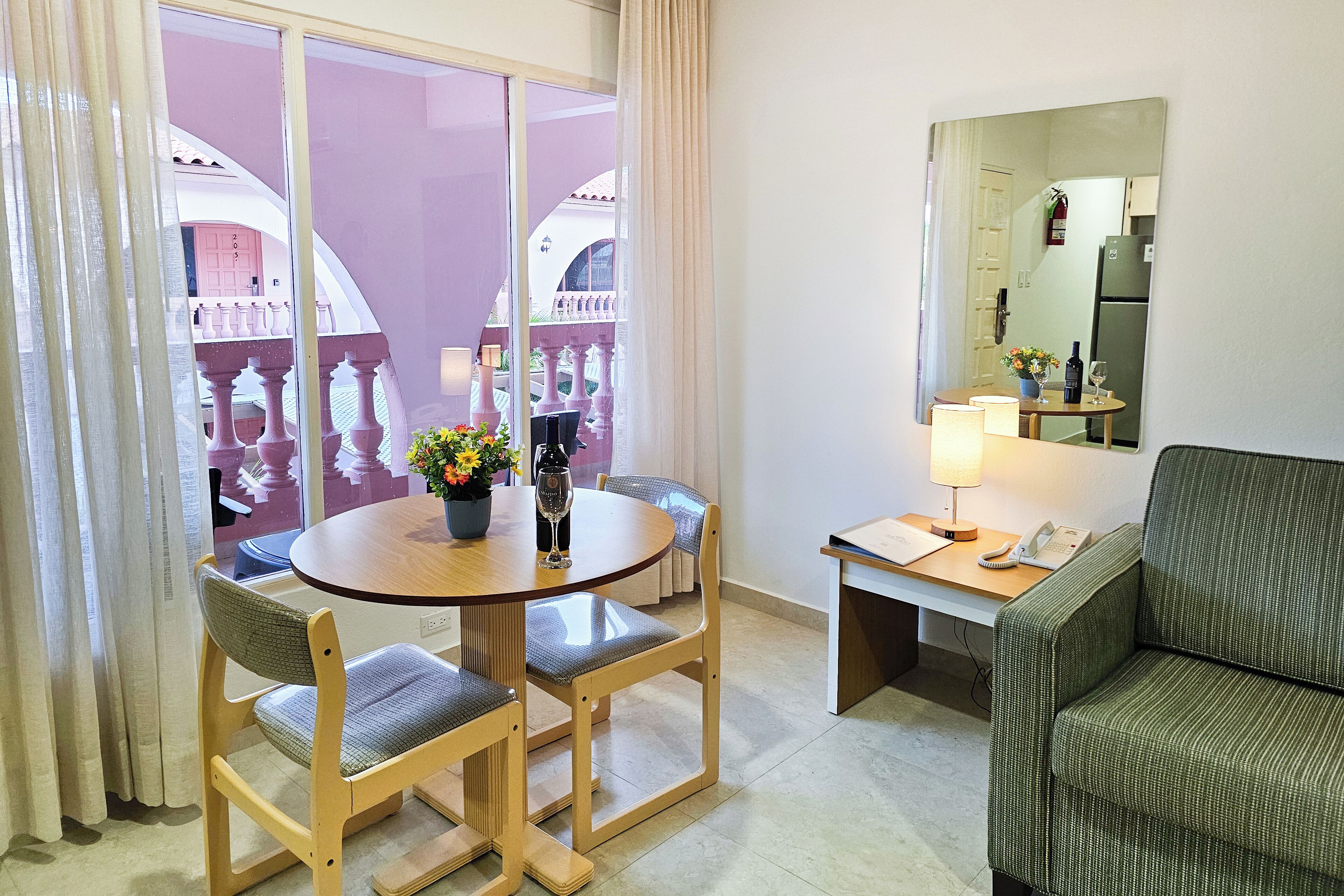 A hotel room with a dining table, two chairs, a sofa, a small desk with a lamp, a telephone, and a large mirror.
