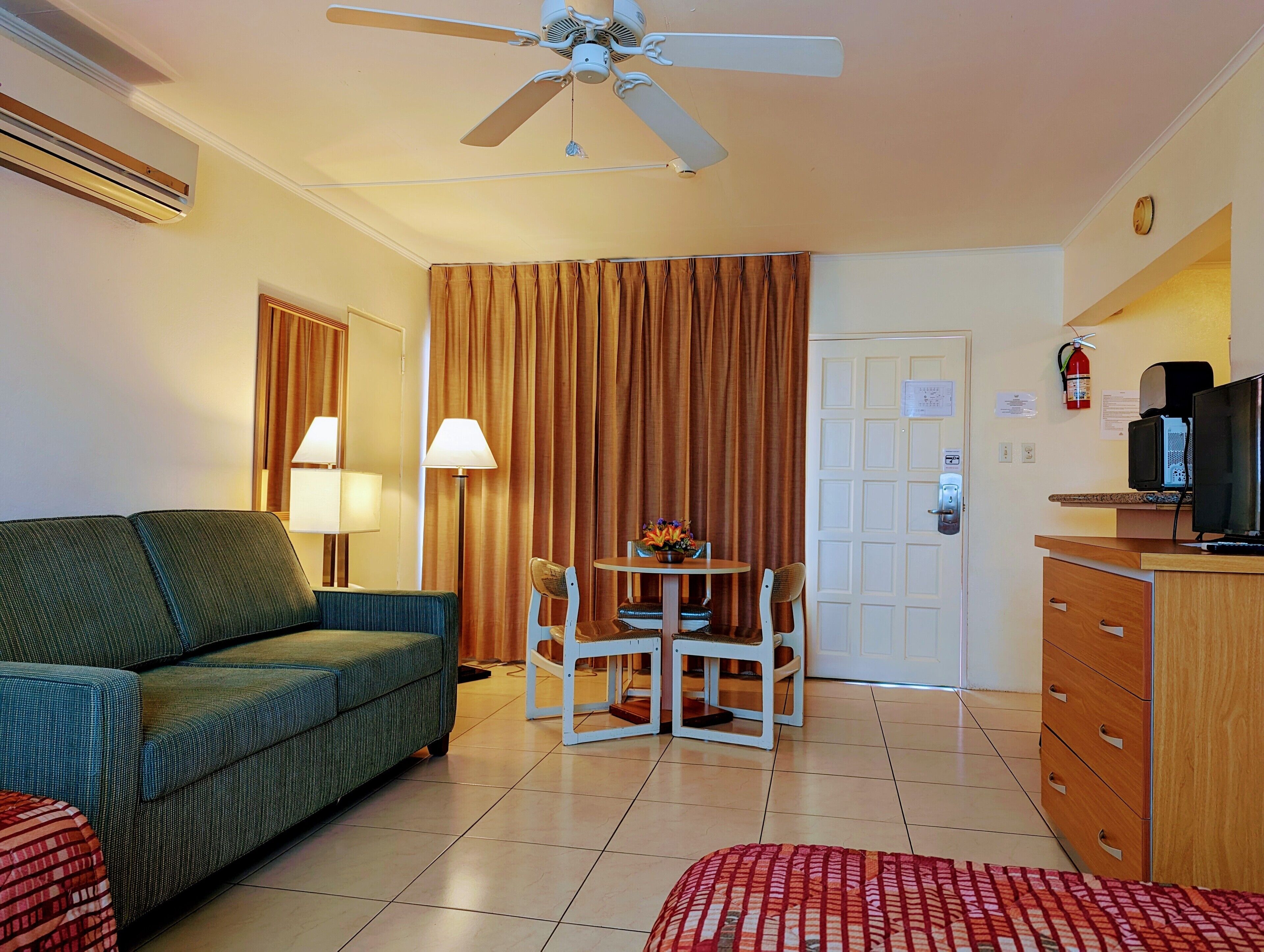Habitación de hotel con sofá verde, una mesa de comedor con sillas, ventilador de techo, televisor y extintor.