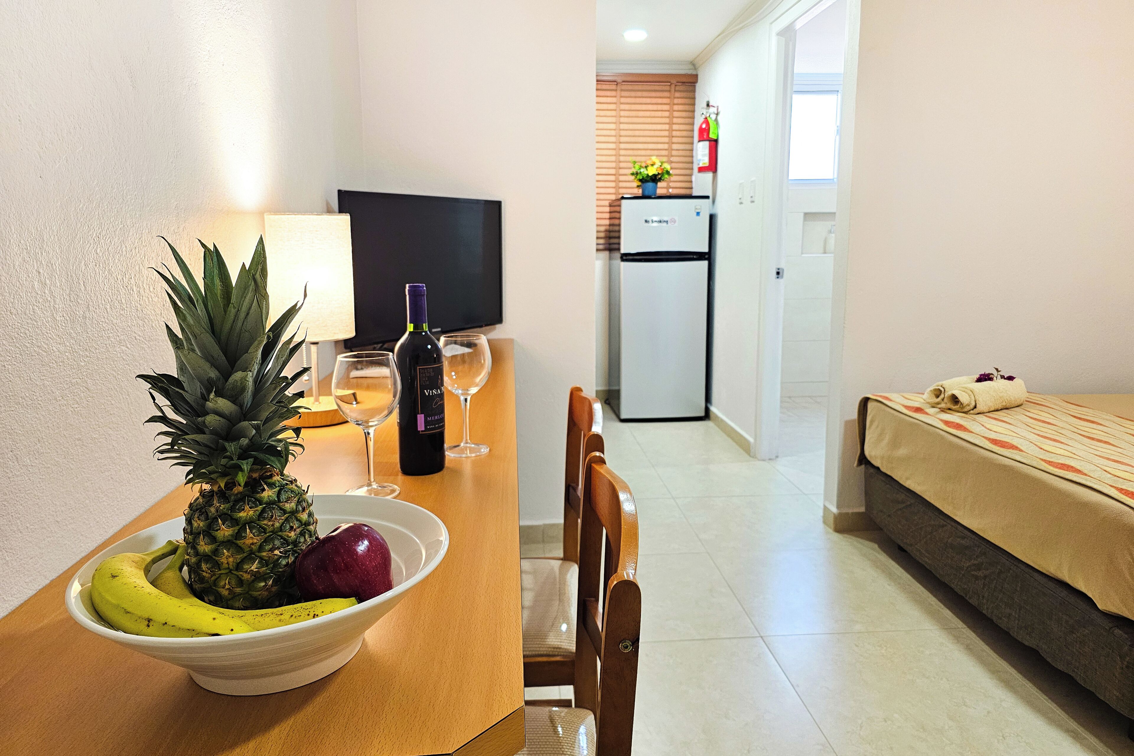 A dining area with a table set for two, a bowl of fruit, a bottle of wine, and a television in the background.