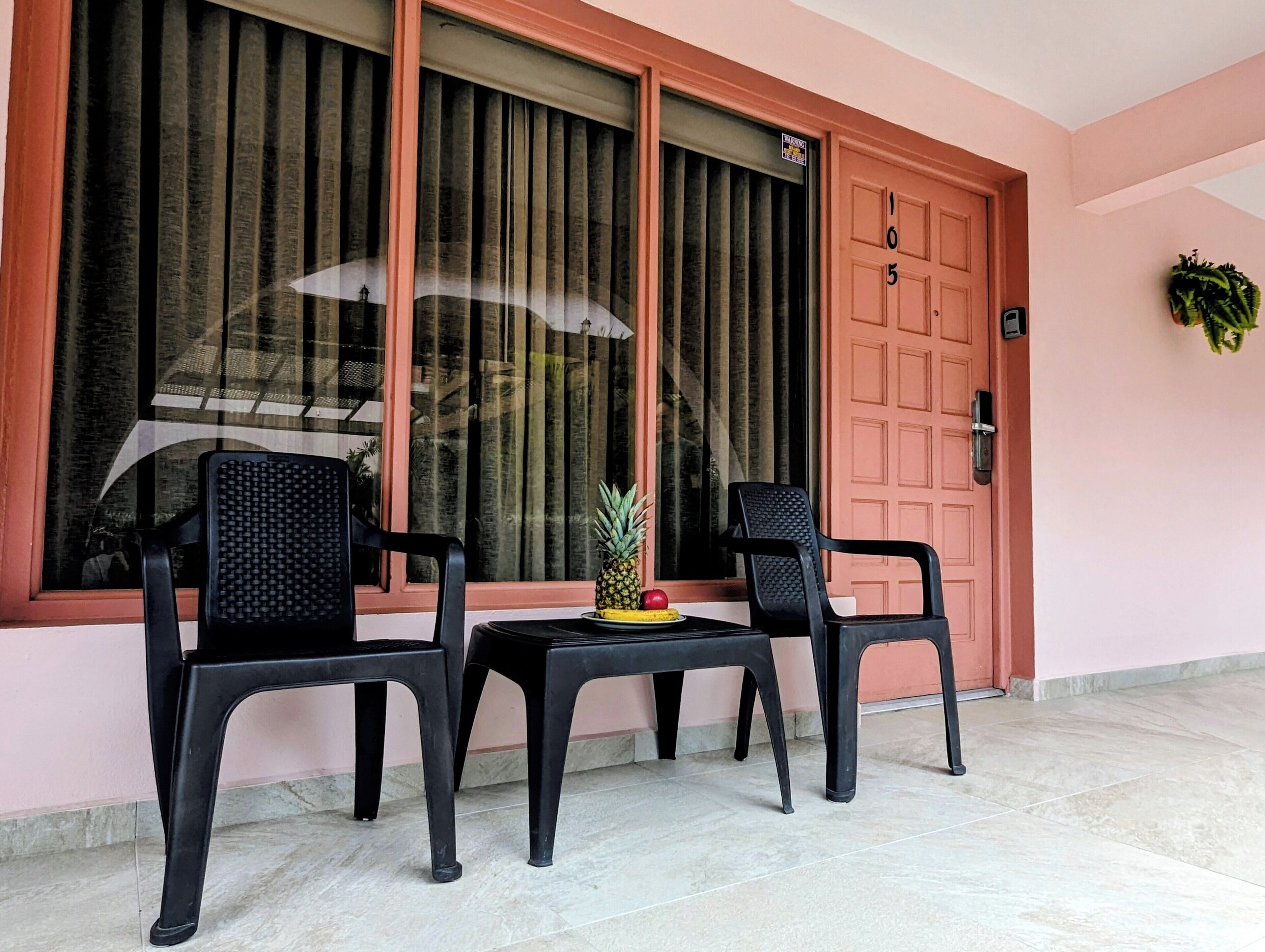 A room with a pink wall, a wooden door with a number, and two black plastic chairs around a small table with fruits.