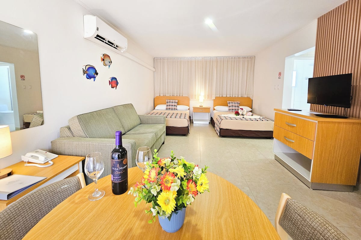 A hotel room with a bed, a desk with a telephone, a TV, and a dining table with a vase of flowers.