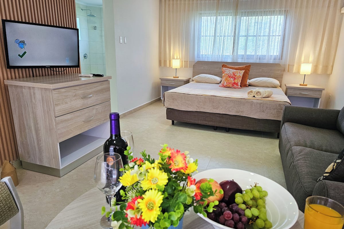A hotel room with a bed, bedside tables, a TV, a dining table with flowers and fruit, and a sofa.