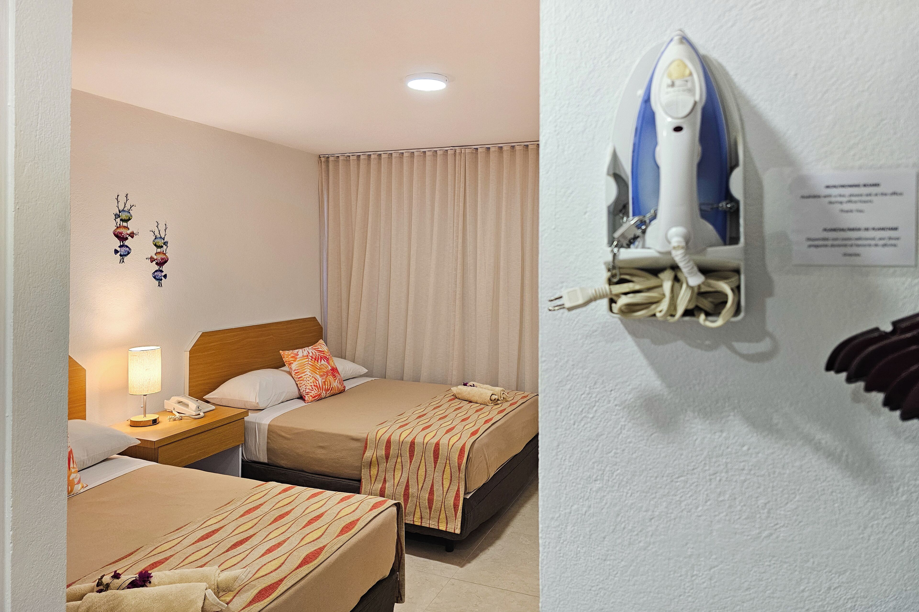 Een hotelkamer met twee bedden, een nachtkastje met een lamp en telefoon, een wandijzer en een gordijn.