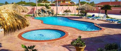 A swimming pool with a thatched umbrella, lounge chairs, and a view of a residential area with palm trees and houses.
