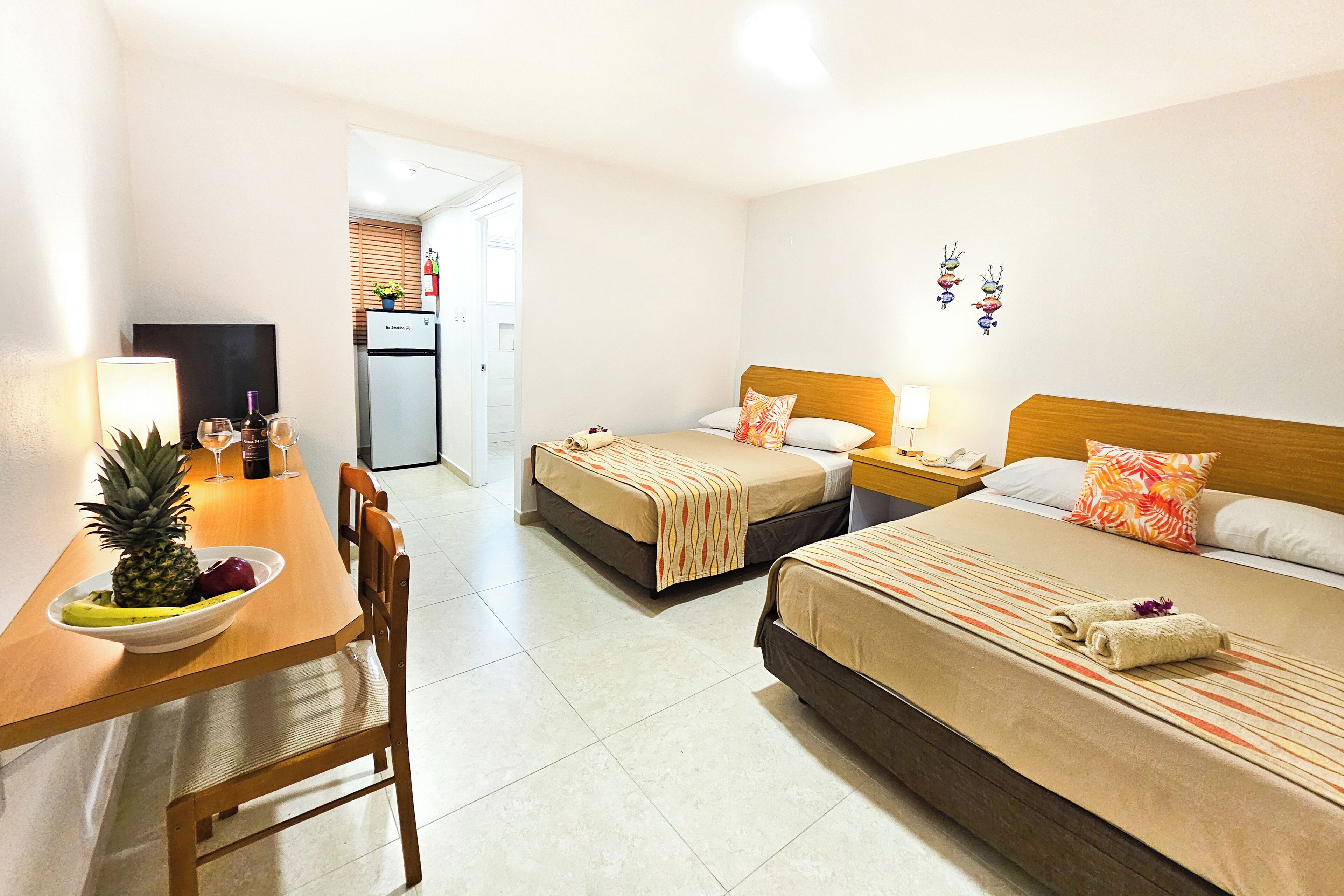A hotel room with two beds, a dining table with fruit, a refrigerator, and a TV.