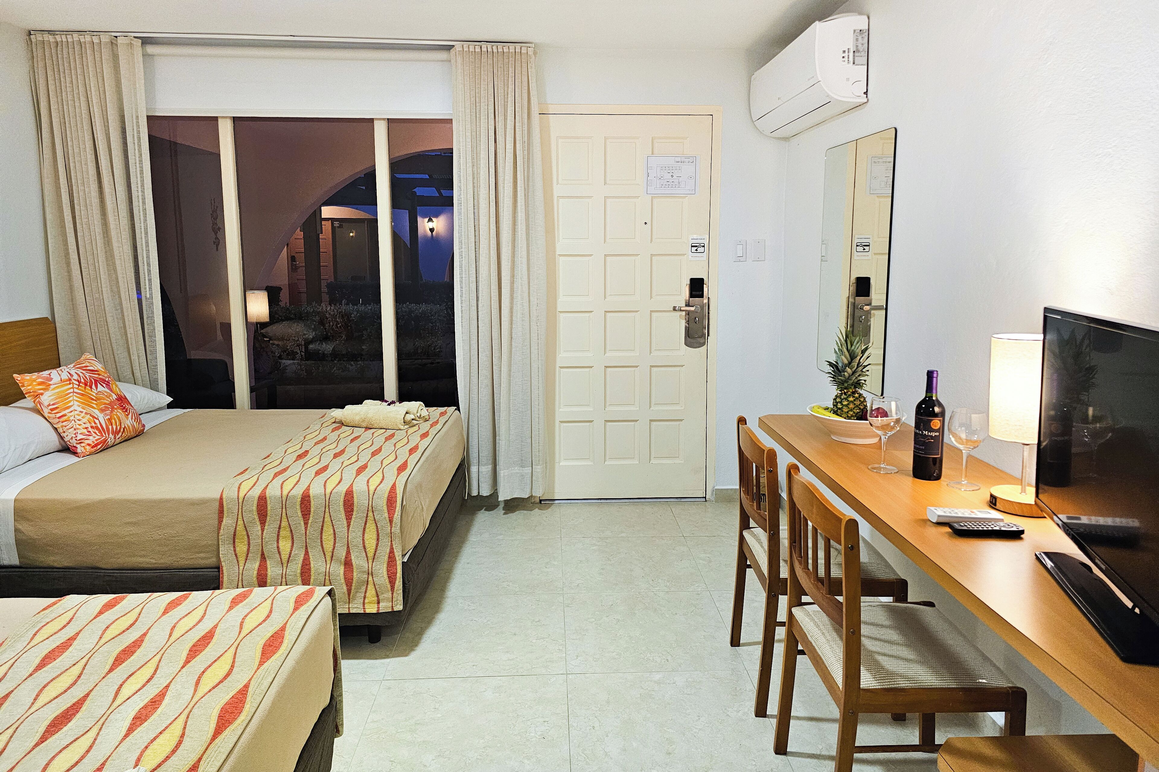 A hotel room with two beds, a desk with a TV, a chair, and a door leading to a balcony.