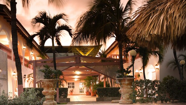 A pool area with palm trees and a building with arched windows and warm lighting. - Sasaki Apartments (Noord)