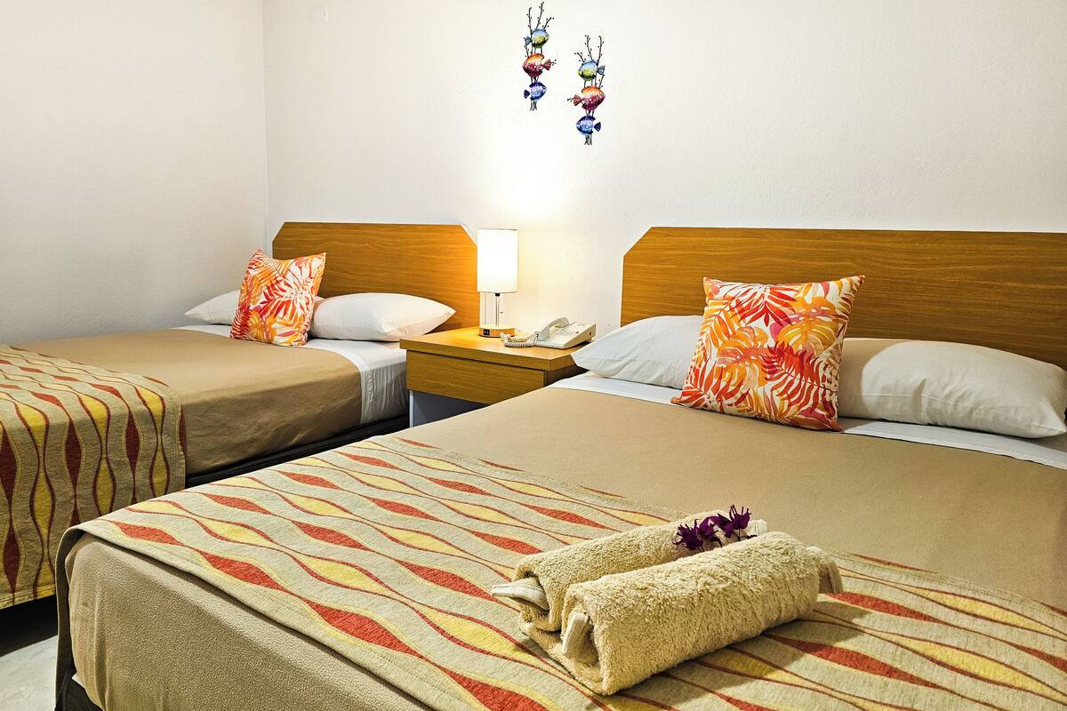 A hotel room with two beds, each with a patterned bedspread and matching pillows. There is a nightstand with a lamp and a telephone between the beds.